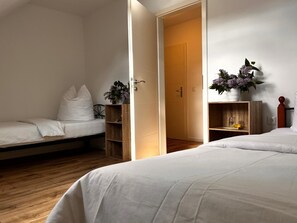 Un dormitorio con dos camas, piso de madera, una puerta que da a otra habitación y una pequeña estantería con un jarrón de flores.
