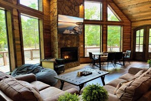 Interior acogedor de una cabaña con chimenea de piedra, paredes de madera y ventanas amplias con vistas al exterior.