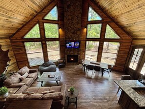 Amplia sala de estar con chimenea de piedra, techo de madera y ventanas grandes que ofrecen vistas al exterior.
