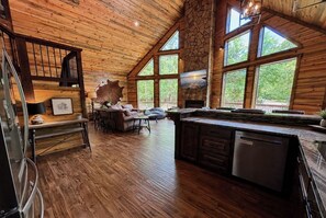 Una habitación amplia con techo de madera, ventanas grandes, chimenea de piedra y comedor con mesa y sillas.