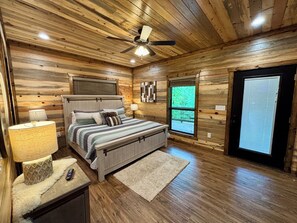 A bedroom with a wooden ceiling, a bed with a headboard, a nightstand, a lamp, a window, and a door.
