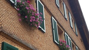 Un edificio de varios pisos con contraventanas verdes, muros de ladrillo y macetas con flores en los alféizares de las ventanas.