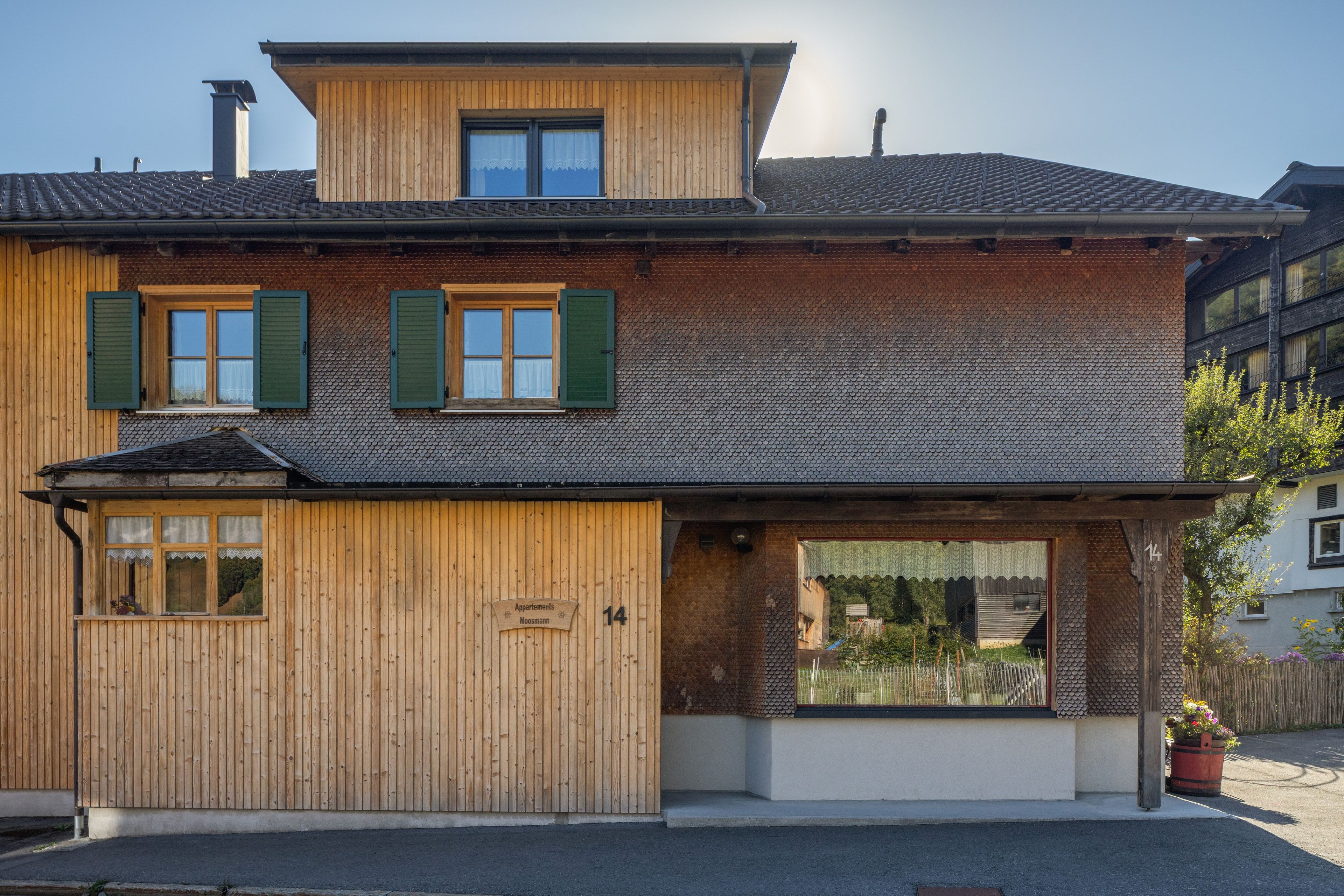 Ein zweistöckiges Holzhaus mit geneigtem Dach, zahlreichen Fenstern mit Fensterläden und der auf der Fassade deutlich sichtbaren Hausnummer 14.
