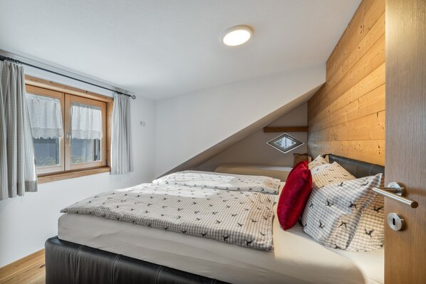 A bedroom with a sloped ceiling, a bed with patterned bedding, a red pillow, and a window with curtains.