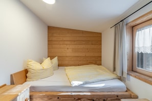 A small, cozy bedroom with a wooden headboard, a single bed with a white mattress and yellow pillows, and a window with a checkered curtain.