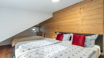 Ein Schlafzimmer mit schrägverdachter Decke, Holzvertäfelung, zwei Betten mit gemusterten Bettbezügen und einem roten Kopfkissen.