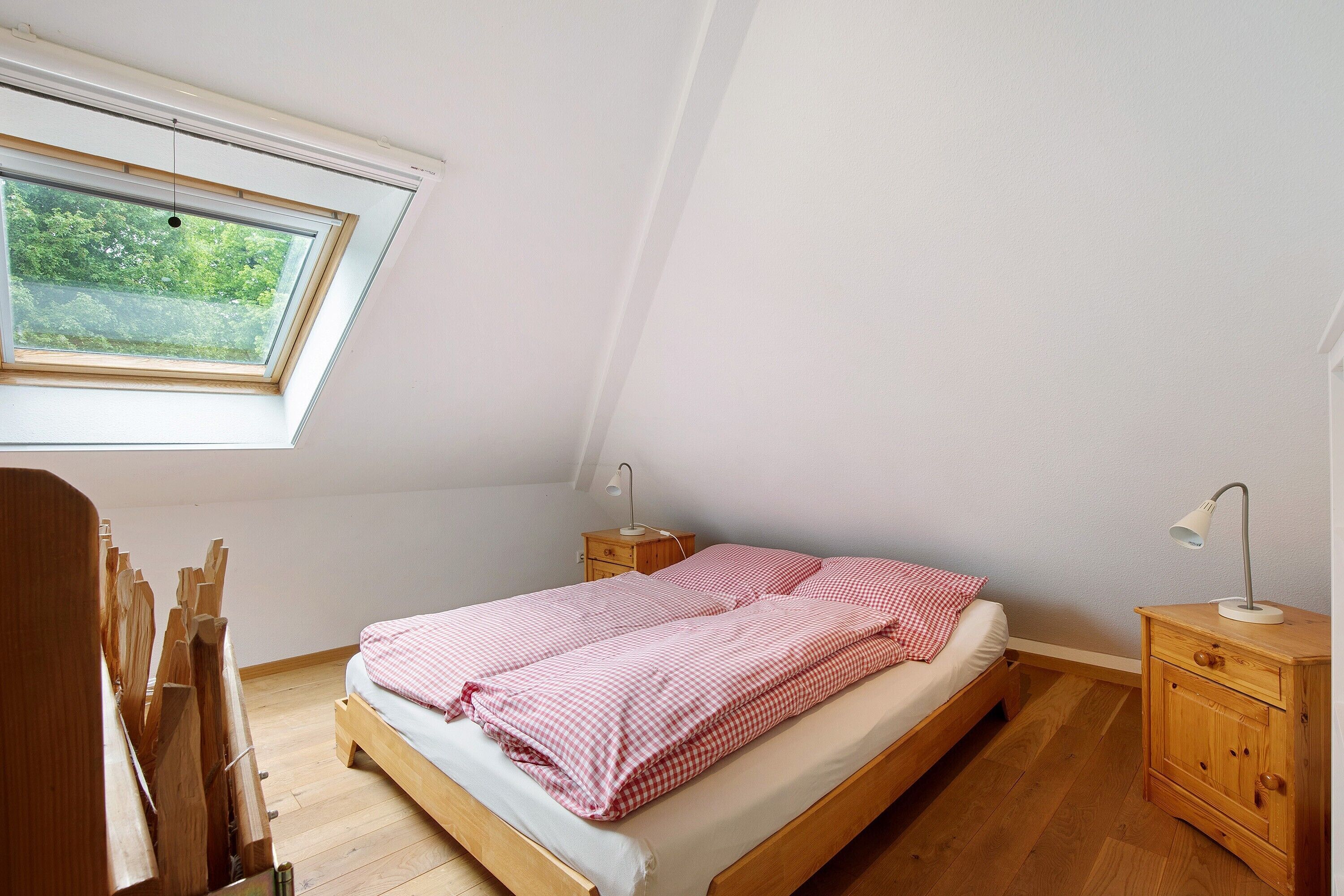 A bedroom with a sloped ceiling, a skylight, a wooden bed, a nightstand, and a desk with a chair.