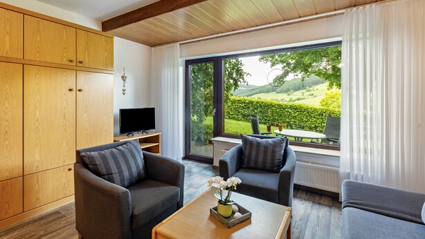 A modern living room with a large wooden wardrobe, two grey sofas, a coffee table with a vase, and a large window offering a view of a garden. - Apartment (Fuchsbau, 30 sqm) with Private Terrace and Wi-Fi (Schmallenberg)