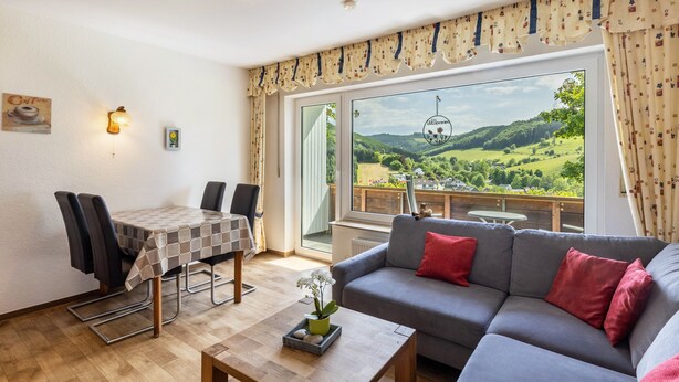 Ein Wohnzimmer mit einer Couch, einem Esstisch mit Stühlen, einem Couchtisch und einem großen Fenster mit Blick auf grüne Hügel.