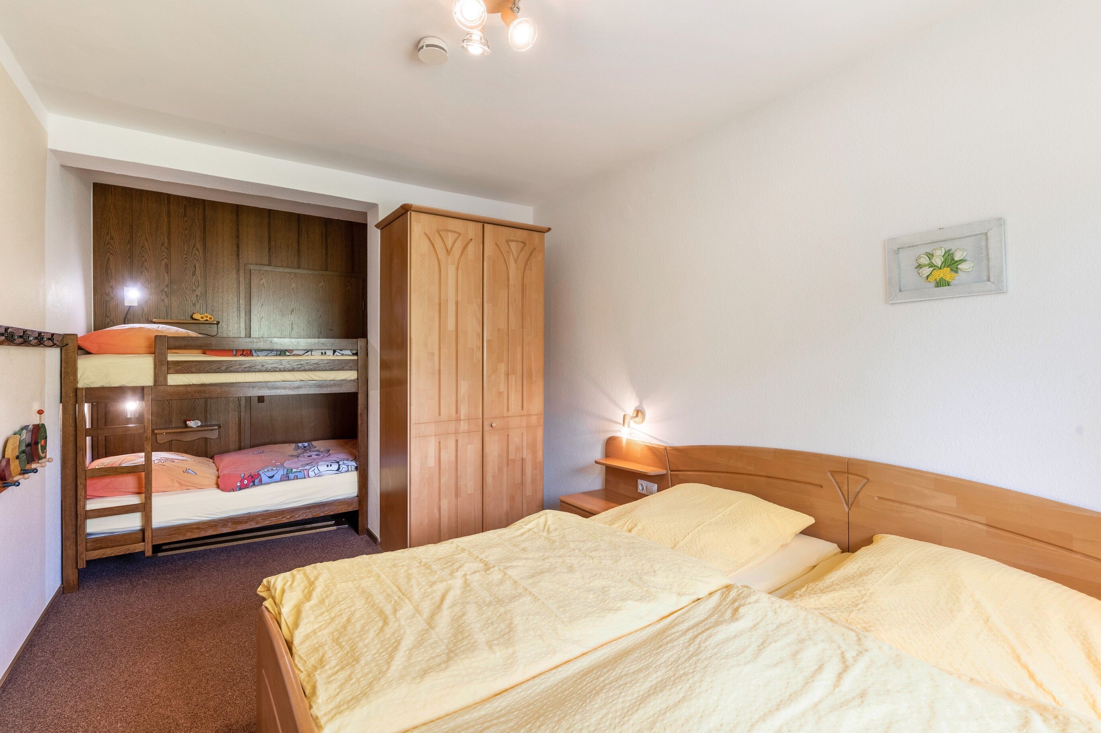 Een kamer met stapelbedden, een houten garderobe, een nachtkastje met een lamp en een klein fotokader aan de muur.