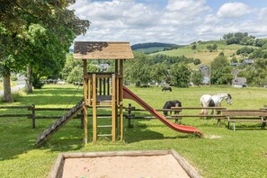 Un terrain de jeux avec un toboggan et une structure d’escalade, entouré d’une zone herbeuse délimitée par une clôture, avec des chevaux qui paissent au loin.