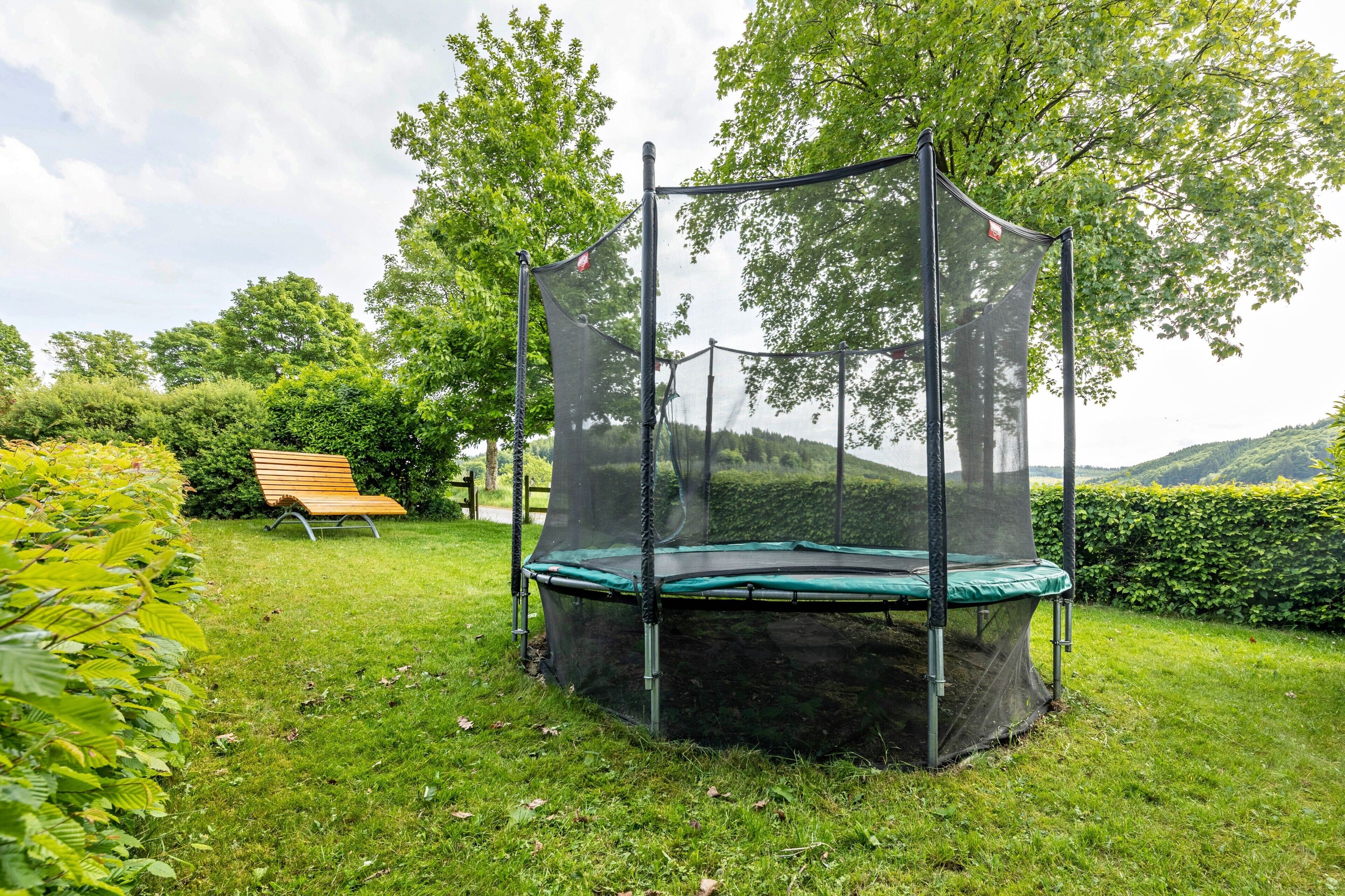 Een trampoline met veiligheidsnet in een grasveld, met een bankje en bomen.