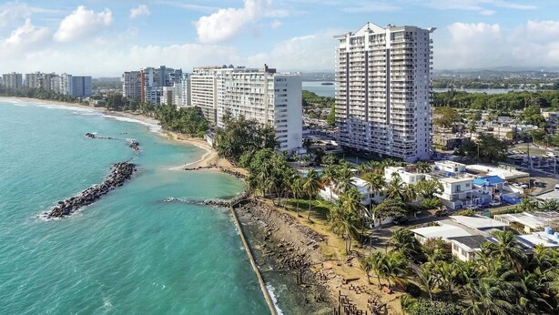 Un paisaje urbano costero con rascacielos, una playa y un rompeolas rocoso.