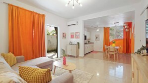 Una sala moderna con un sofá, una mesa de centro y cortinas anaranjadas. Se ve un área de cocina con una mesa de comedor y sillas a través de un pasillo abierto.