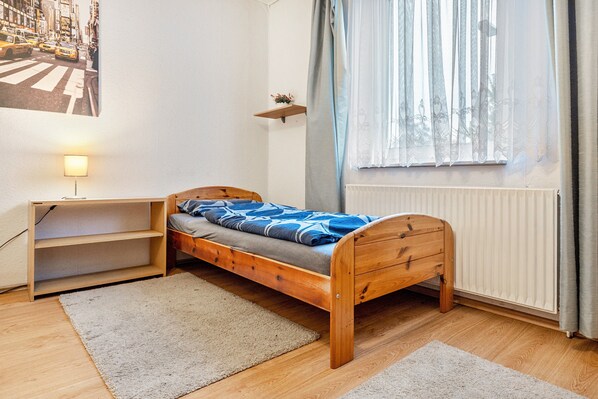A small, single-bed room with a wooden frame, a bedside table, a shelf, a window with sheer curtains, and a radiator.