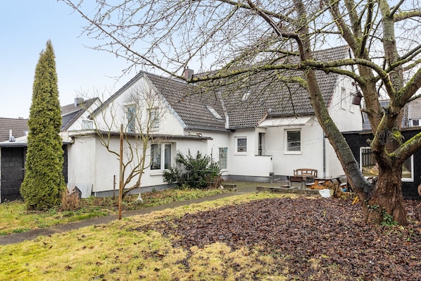 Une maison d’habitation avec un toit en pente, un chemin menant à la porte d’entrée et une cour où se trouve un tas de feuilles.