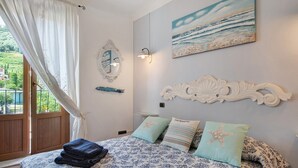 A bedroom with a bed, a window with curtains, a painting of the sea, and a decorative wall piece.