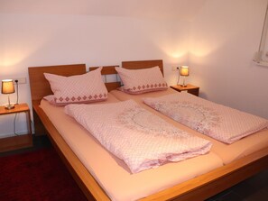 Uma cama de casal com dois mesaninhos de cabeceira, cada um com uma luminária, num quarto com paredes brancas e tapete vermelho.