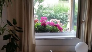 Een kamer met een raam dat uitkijkt op een tuin, een nachtkastje met een lamp en een bed met een blauwe deken.