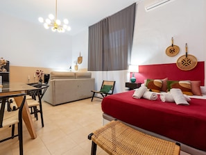 Una habitación de hotel con cama, una mesa de comedor, sofá, sillón, lámpara y aire acondicionado instalado en la pared.
