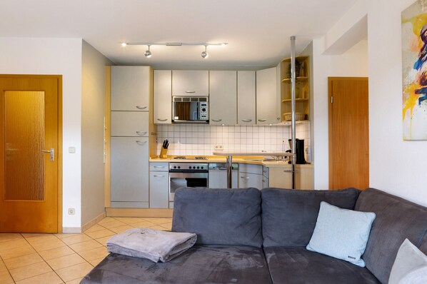 Un espace de vie compact comprenant un coin cuisine, un canapé gris et une porte en bois.