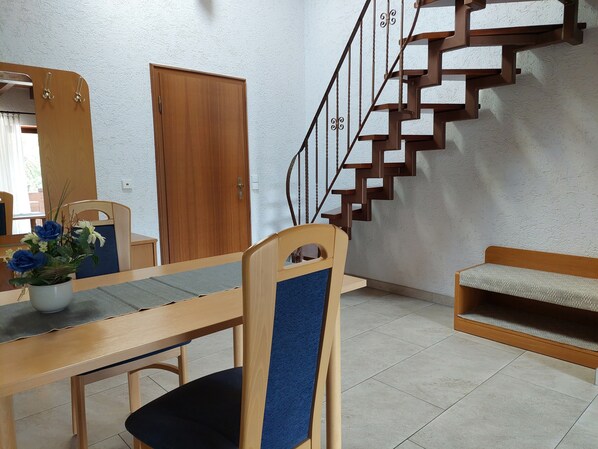 Un comedor con una mesa de madera, sillas, un jarrón con flores, una escalera con barandilla y una puerta.