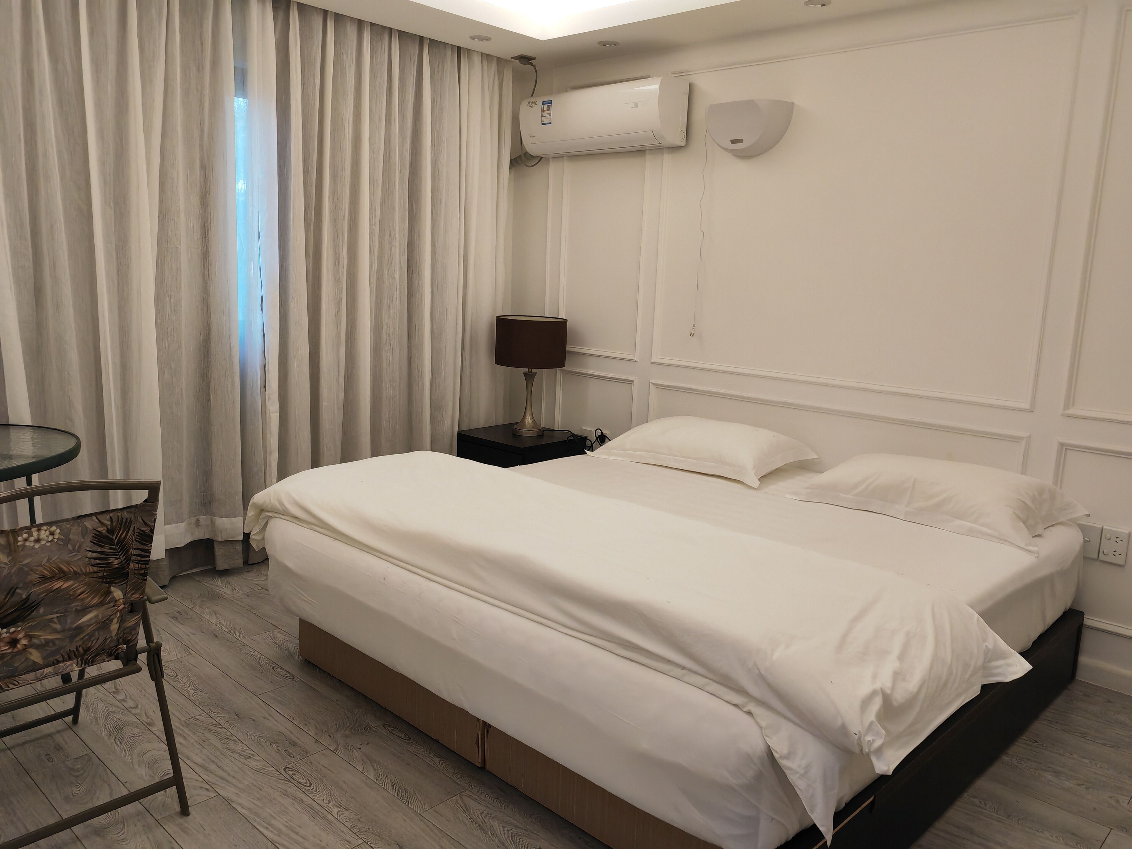 A hotel room with a bed, a nightstand, a lamp, a chair, and an air conditioning unit.