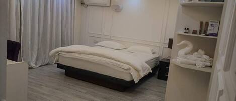 A modern bedroom with a large bed, a bedside table, a wardrobe, and a shelf with towels and toiletries.