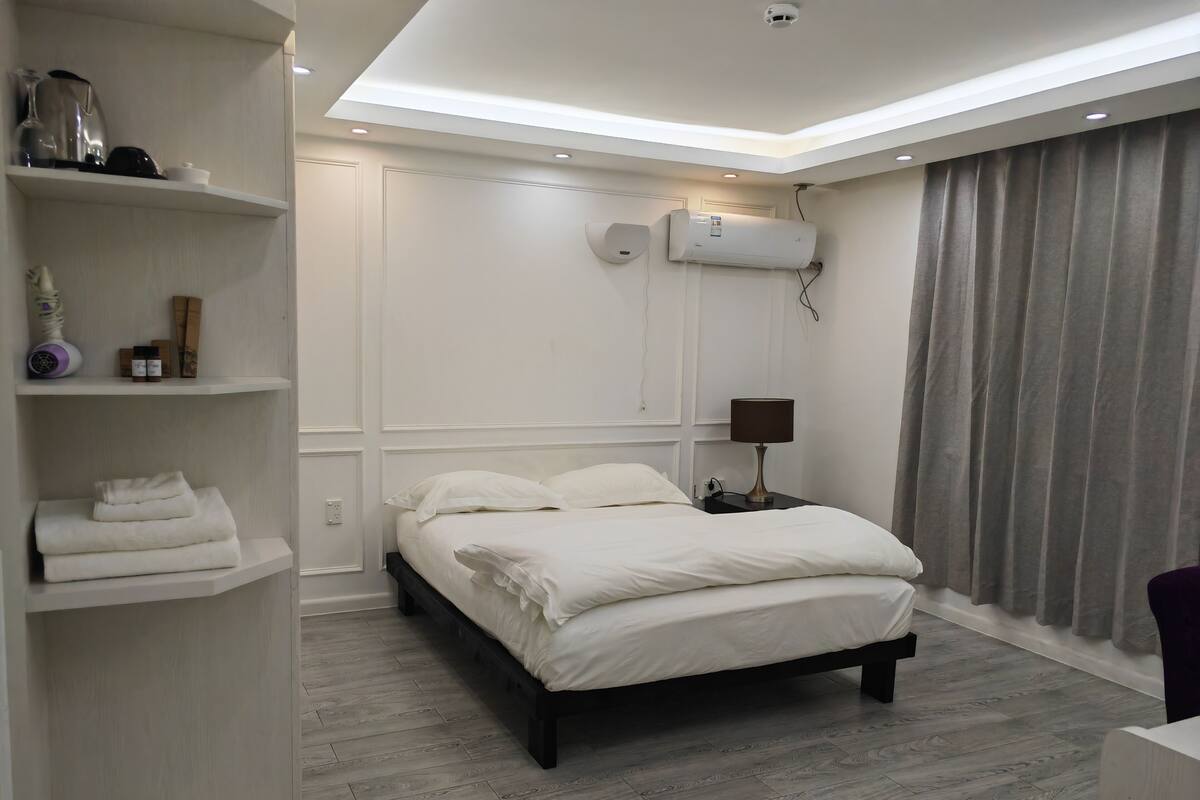 Un dormitorio moderno con una cama, una mesita de noche, una lámpara, un estante con toallas, un aire acondicionado instalado en la pared y una ventana con cortinas.