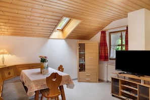 A cozy room with a wooden ceiling, a skylight, a dining table with chairs, a TV on a stand, and a cabinet with glass doors.