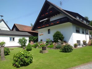 A traditional building with a sloped roof, a balcony with flowers, and a well-maintained garden with various plants and a lawn.