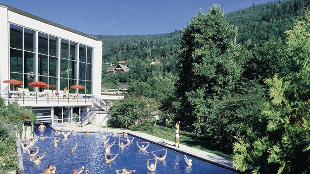 A swimming pool with people exercising, surrounded by a building with large windows and a forested hillside.