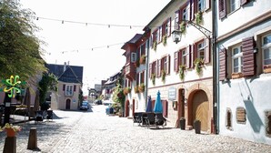 Una calle adoquinada flanqueada por edificios tradicionales europeos, algunos con macetas de flores y mesas al aire libre.