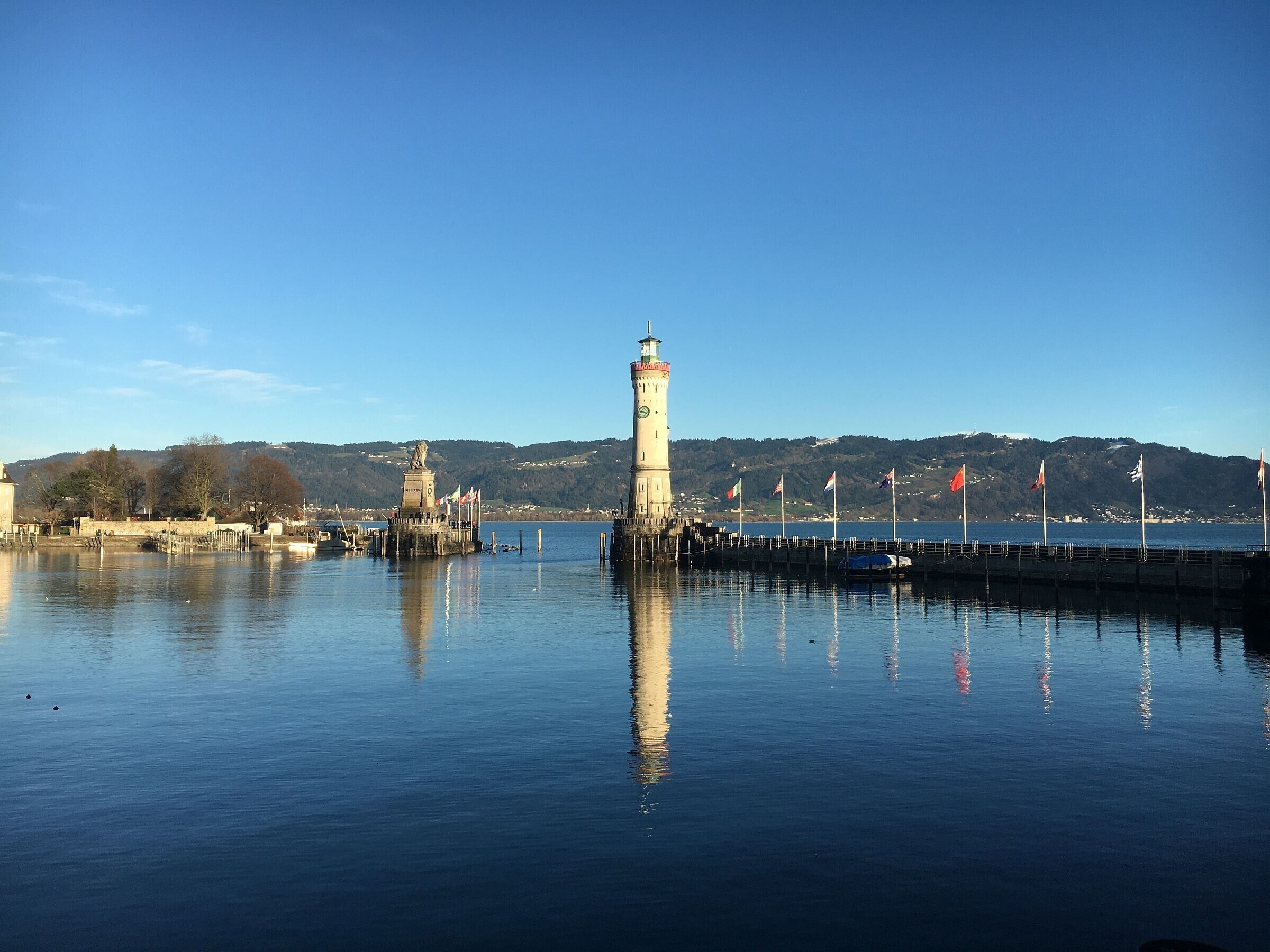 Una orilla tranquila con un faro destacado, un muelle que se adentra en el agua y colinas al fondo.