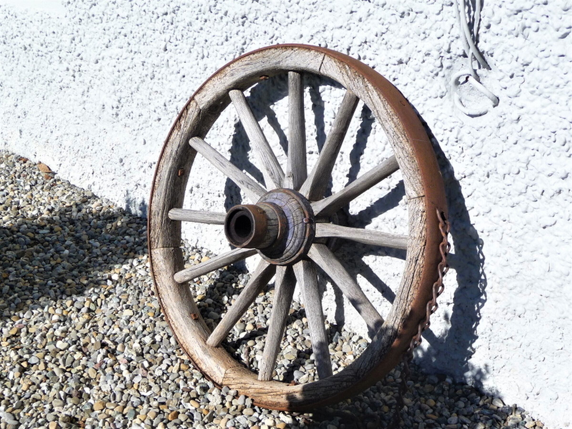 A wooden wheel with spokes and a central hub, lying on a gravel surface against a white wall.