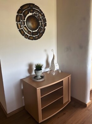 Een hoekje met een houten consoles, een kamerplant, een decoratief metalen wandobject en een witte plank met een spiraalvormig ontwerp.