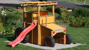 Estructura de juego con forma de vehículo de construcción amarillo, que incluye un tobogán, rodeada por un jardín con flores y una valla.