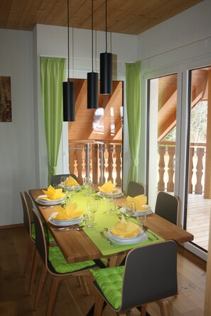 Un coin repas avec une table en bois dressée pour six personnes, des rideaux verts et des lampes suspendues.