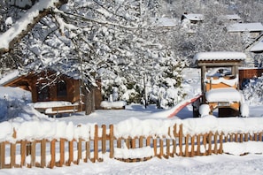 Uma cabana de madeira com telhado coberto de neve, rodeada por árvores carregadas de neve e uma cerca de madeira.