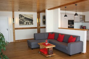 Een moderne woonkamer met een grijze bank, rode kussens, een houten salontafel en een keuken met kasten en een spoelbak.