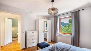 A bedroom with a bed, a wardrobe, a small table, and a window with a view of greenery.