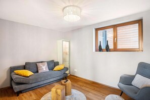 A modern living room with a grey sofa, wooden coffee table, and a grey armchair. A large window with wooden shutters and bottles on the sill is visible.