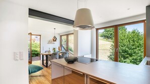 A modern kitchen with a large island, a dining area with a table and chairs, and large windows offering a view of greenery.
