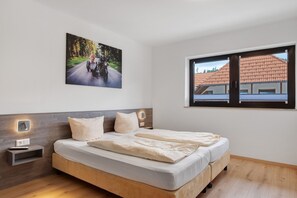 A bedroom with a bed, a nightstand, a wall-mounted light, a picture of a motorcycle on a road, and a window with a view of buildings.