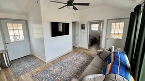 Ein geräumiges Wohnzimmer mit Deckenventilator, einem großen Flachbildfernseher an der Wand, einem beigefarbenen Sofa mit farbenfroher Decke und einem gemusterten Teppich auf Holzdielen.