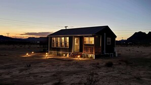Eine kleine Holzhütte mit Veranda, eingebettet in eine Wüstenlandschaft im Abendlicht.
