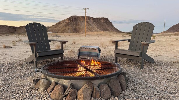 Zwei Adirondack-Stühle sind einem Metall-Feuerkorb mit Steinen gegenüber, eingebettet in eine felsige Outdoor-Umgebung mit einem Berg im Hintergrund.