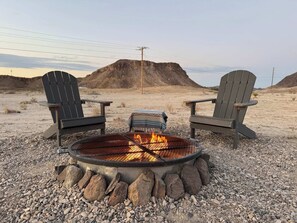 Two Adirondack chairs facing a metal fire pit with rocks around it, set in a rocky outdoor area with a mountain in the background. - MJ's Cabin. Private cabin just 3.5 miles from BBNP (Terlingua)
