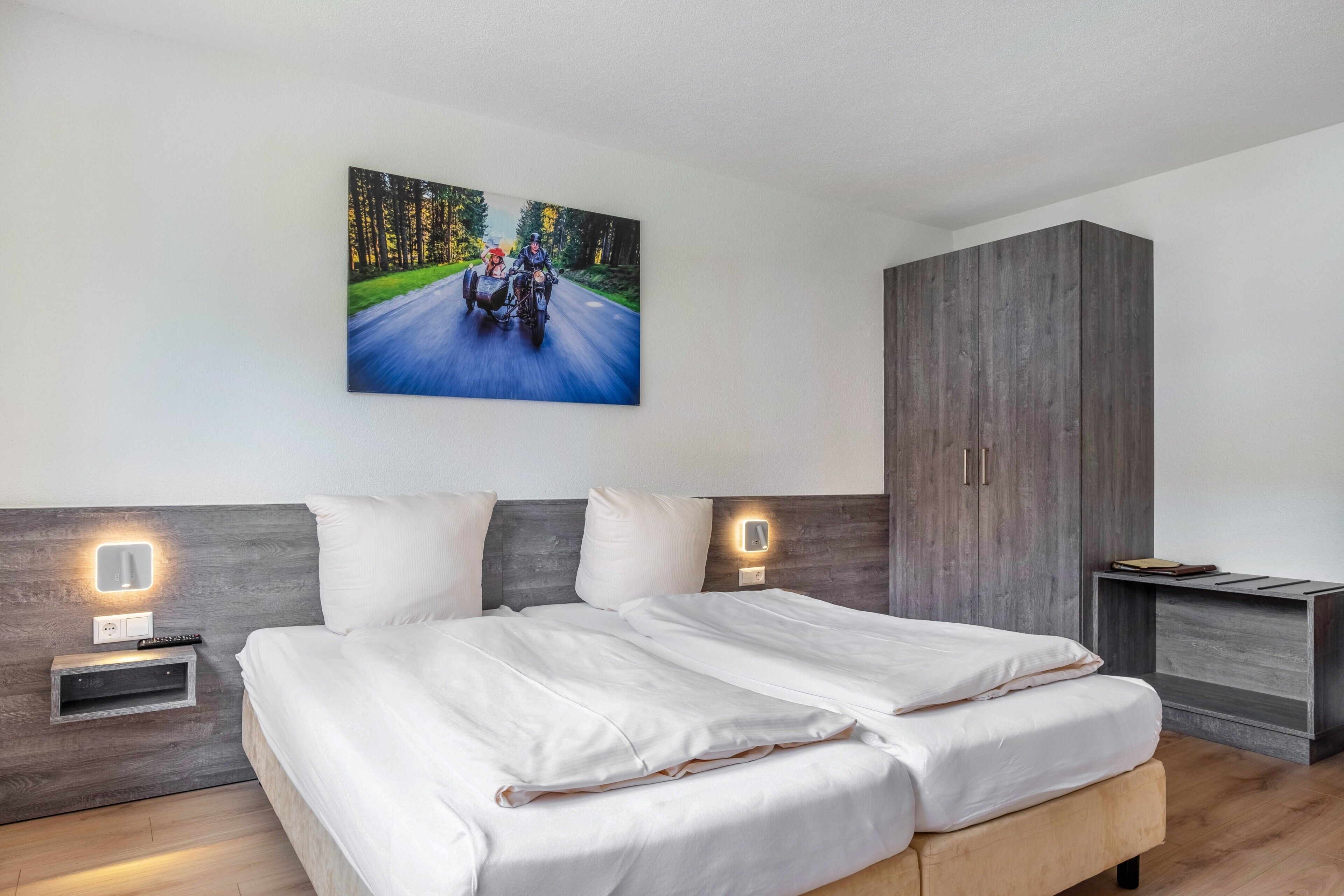 Quarto moderno com cama grande, mesa de cabeceira com abajur, guarda-roupa e um quadro de uma motocicleta em uma estrada.