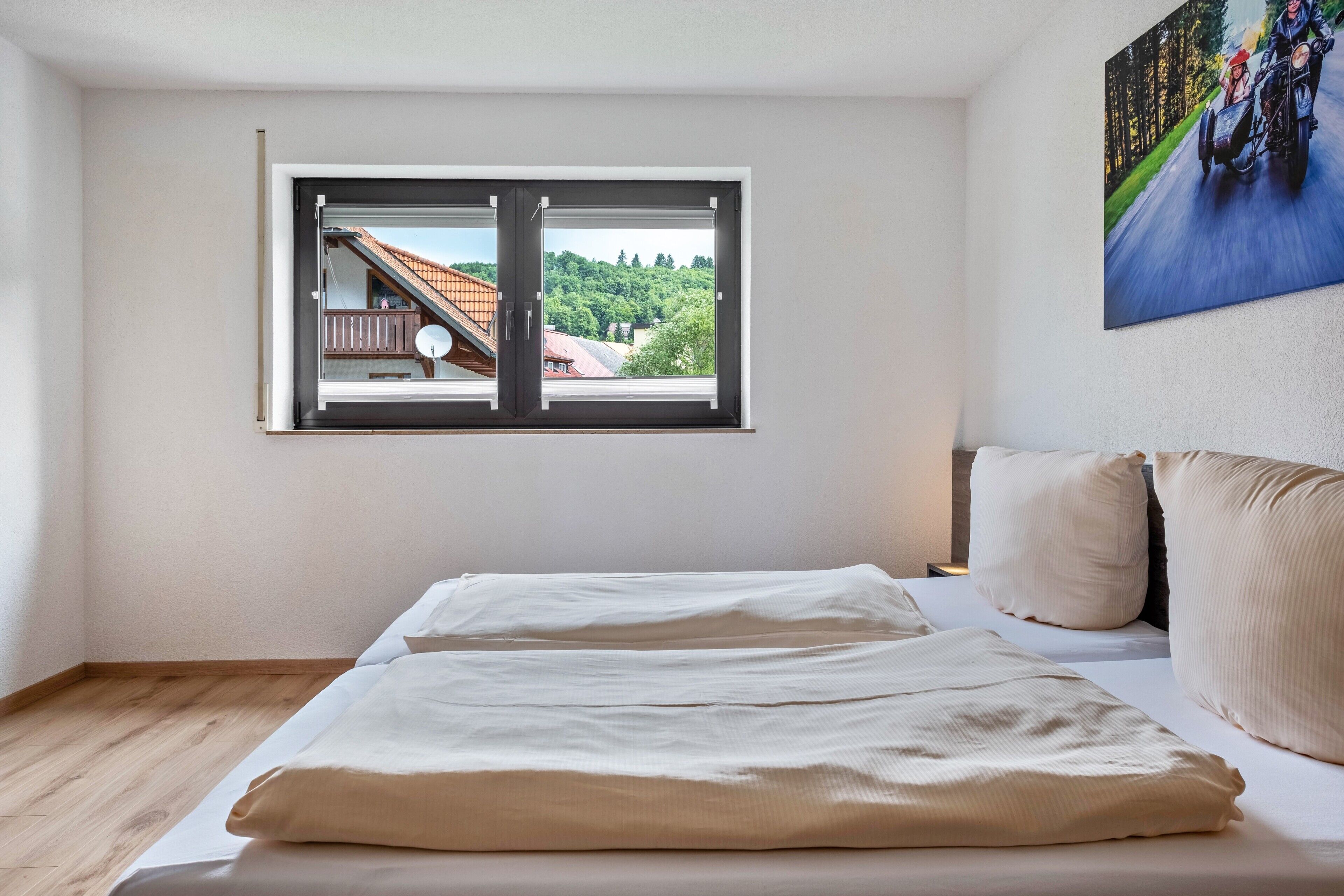 Un dormitorio con cama, almohadas, una ventana que ofrece vistas a un edificio y árboles, y un cuadro en la pared.